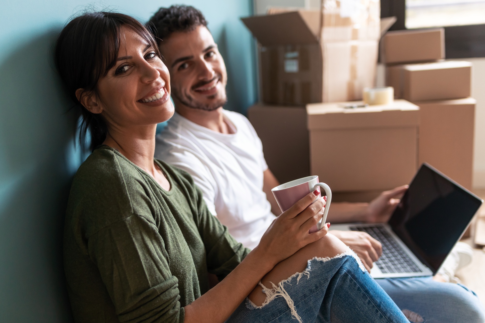 Couple searching ideas with laptop while drinking coffee looking at camera sitting on the floor.